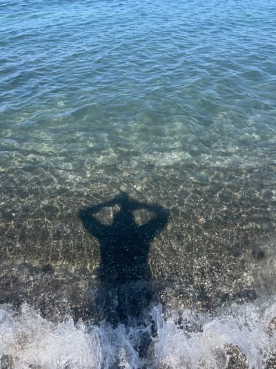 The image depicts a clear, shallow body of water with sunlight refracting through it, illuminating the pebbled seabed. A shadow of a person is visible in the water, with their hands raised to their head. The shadow is cast from the right side of the frame and extends towards the left. The water's surface exhibits gentle ripples. In the foreground, white foamy waves are breaking on the shore. The location is the Black Sea coast in Shekvetili, Georgia.
