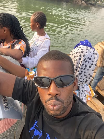This media file captures a selfie taken by a man on a boat, surrounded by several other people. The man in the foreground is wearing black sunglasses, which reflect the bright daylight and parts of the boat's interior, and a black t-shirt with a blue embroidered logo on the left chest. He has a short beard and mustache.
Behind him, to his right, a woman with braided hair is visible, wearing a white long-sleeved shirt with green lettering, and a gold chain around her neck. Further back and to her right, another individual with braids wears a colorful patterned top. In the background, partially obscured, is a person facing away from the camera, dressed in a white and black star-patterned garment over a blue piece of clothing. A light brown suitcase and blue jeans are also visible, suggesting travel.
The setting is a body of water, likely a river or lake, with visible ripples across its surface, indicating movement or gentle currents. The water appears green-tinted from reflections. Portions of a wooden boat structure are visible, and on a grey surface in the boat's lower right, the reversed text "Toyo" can be read. The overall scene suggests a bright, sunny day, indicating clear weather. The activity appears to be a boat trip or journey. This scene is situated in Gburuku, Nigeria.