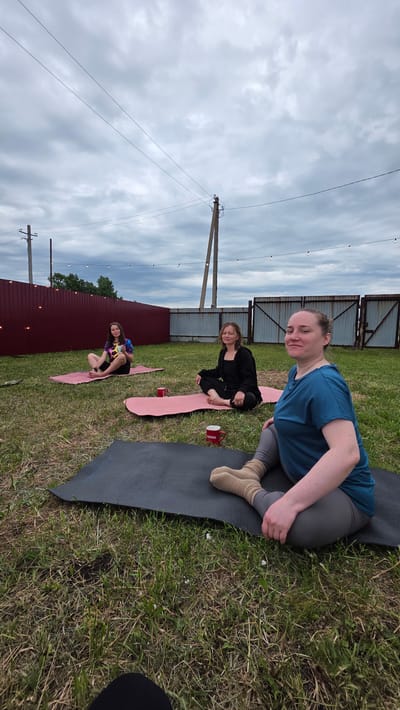 In Купоярово, Russia, three women are practicing yoga on a grassy backyard. Two women are seated on pink yoga mats, while the third is on a black yoga mat in the foreground. The woman in the foreground is wearing a teal t-shirt and grey leggings, and is in a seated yoga pose. The other two women are also seated in meditative poses. Two red mugs are visible on the yoga mats. A maroon fence runs along the background, with a metal gate to the right. Power lines and utility poles are visible in the cloudy sky.