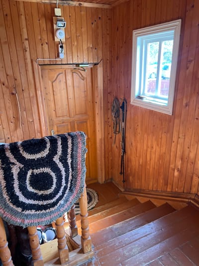 The image captures an interior view, likely within a residence, in Чернышово, Russia. The space is characterized by extensive use of wood, evidenced by the wooden walls, ceiling, and staircase. A rectangular window is situated on the ceiling, allowing natural light into the room. Adjacent to the window, on the ceiling, there's a pair of ski poles and associated straps. On the wall there's a light switch and a door with an ornate handle. A circular rug featuring a black and white concentric design is prominently displayed in the center, partially obscuring the view of a wooden staircase with a matching rug at the top. The stairs are painted brown.
