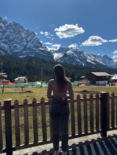 A medium, landscape-oriented shot captures a woman looking at a mountain view from a wooden platform or balcony. The scene is likely set in Sappada, Italy, given the mountainous terrain typical of the region.
The woman, positioned in the center of the frame, stands with her back to the viewer. She has long brown hair and wears a light gray long-sleeved shirt and dark gray jeans. Her hands seem to rest on the wooden fence or railing in front of her.
Behind the woman, a valley stretches out, featuring a mix of evergreen trees and grassy fields. Several buildings dot the landscape, including a house with a brown roof and white walls. The mountain range dominates the background, its rocky peaks partially covered with snow. The sky is bright blue with scattered white clouds.
In the foreground, the wooden fence runs horizontally across the frame, parallel to the woman's body. A section of the platform is visible, constructed from a metal grating. The lighting suggests a sunny day, casting shadows and creating highlights on the objects and surfaces.