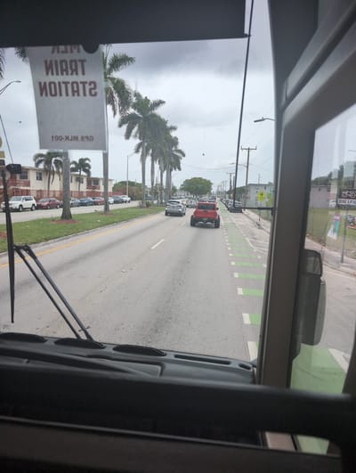 The image shows a view from inside a bus looking out the front windshield and side window. The bus is driving on a street in Opa-locka, Florida. Palm trees line the street, and there are several cars and buildings visible. A sign hangs from the windshield that reads "TRAIN STATION" upside down. A red truck is driving in front of the bus, and there is a bike lane marked with green paint on the right side of the road. The sky is cloudy.