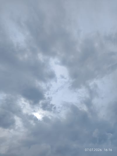 The image displays a cloudy sky over Konovalovo, Russia, on July 7, 2026, at 16:16. The sky is filled with various shades of gray clouds, with patches of lighter sky visible through some breaks. The sun is partially obscured by the clouds, creating a soft illumination. No people, subjects, or objects are clearly identifiable in the scene. The environment is solely characterized by the atmospheric conditions.