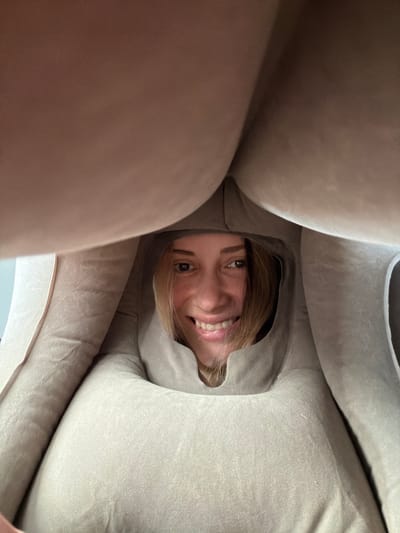 A young, fair-skinned woman with brown eyes and blonde hair smiles directly at the camera from within a soft, beige or light brown fabric enclosure. The fabric is bunched around her face, creating a tunnel-like perspective. The image is taken from a low angle looking upward at the woman. The environment appears to be indoors. There is no information to suggest the image was taken in совхоза "Воронежский", Russia.