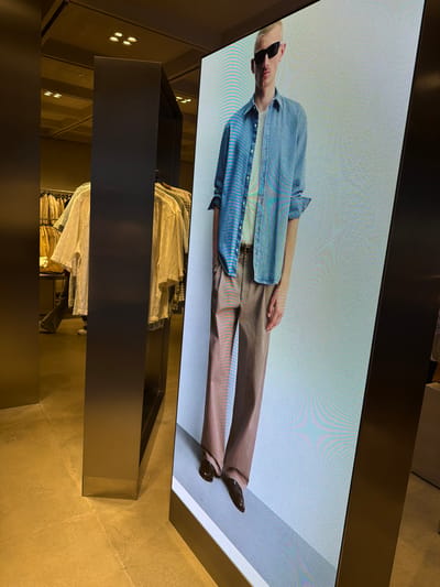 The image shows the interior of a clothing store in Qinhuai District, China. A large, angled LED screen displays a full-body advertisement of a light-skinned male model wearing sunglasses, a light blue denim shirt, a white t-shirt, and tan trousers. The model stands with his hands in his pockets. In the background, clothing racks with various garments are visible. A dark, metallic pillar stands to the left of the screen, and the floor appears to be a polished, light-colored tile. The lighting is dim, with spotlights visible on the ceiling.