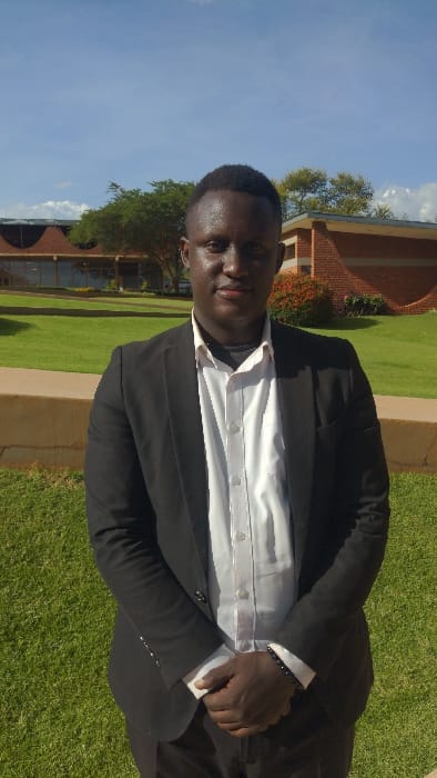 This image captures a young man posing outdoors in Karatu, Tanzania, during daytime. The man, with dark skin and short, dark hair, is dressed in a black suit jacket over a white button-down shirt. A dark, beaded bracelet adorns his left wrist. He stands on a neatly manicured green lawn, looking directly at the camera with a neutral to slightly serious expression. A subtle sheen is visible on his skin, suggesting bright sunlight or warmth.
The background features a well-maintained outdoor environment. Light brown paved pathways or borders segment areas of green grass. Further back, various trees and bushes provide greenery. Prominent brick buildings with reddish-brown roofs are visible, characteristic of an institutional or campus setting. The sky is bright blue with a few scattered white clouds, indicating clear and sunny weather. The strong illumination and distinct shadows confirm it is mid-morning or afternoon. No text is visible in the image. The overall scene suggests a formal or semi-formal portrait.