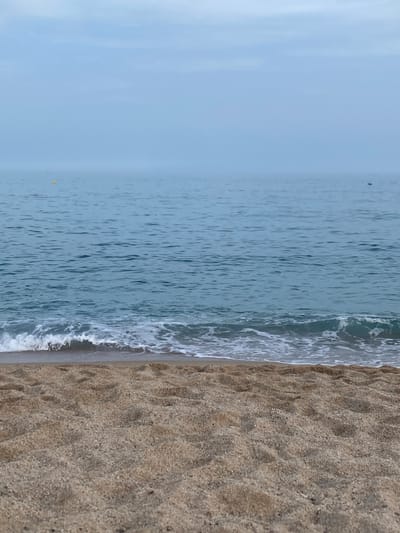 This is a photograph of the beach in Vilassar de Mar, Spain. The foreground is a sandy beach, with the sand appearing light brown and slightly damp. The texture of the sand shows small pebbles and indentations. In the midground, gentle waves with white foam are washing onto the shore, creating a line of surf. The water itself is a calm, blue-green color, with a slight ripple pattern. In the distance, the sea meets the sky in a hazy horizon. The sky is overcast and a pale blue-gray color, with faint clouds. There are two small, dark objects visible on the water in the far distance, which could be boats. One yellow buoy is visible in the far left of the frame. The overall atmosphere is muted and serene.