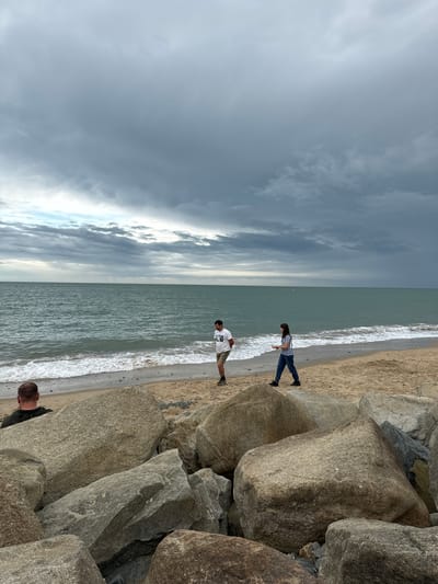 A group of people are on a beach in Courtown, Ireland, with the sea in the background under a cloudy sky. The foreground is dominated by large rocks. A man with a short haircut is sitting on the rocks facing away from the camera. Further back, a man in a white t-shirt and shorts walks along the sand towards the sea. To his right, a woman in a striped t-shirt and dark pants walks along the sand. The sea is choppy with waves crashing on the shore. The sky is filled with dark, heavy clouds, suggesting an overcast or stormy day.