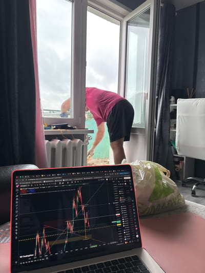 This media file captures a scene indoors, with a clear view of an open window leading to an outdoor environment. The primary subjects are a person and a laptop. The person, seen from the back, is wearing a pink t-shirt and dark shorts and appears to be reaching out or working near the window, possibly engaged in some repair or adjustment. A white plastic bag with green markings rests on a surface to the right of the person.
The setting seems to be a room with dark curtains. The laptop, positioned on a pink surface, displays a financial trading chart, specifically showing Bitcoin (BTCUSDT) data from TradingView. The chart indicates various price movements and indicators. Visible text on the laptop screen includes website URLs, cryptocurrency names like BTCUSDT, and numerical data.
The activity suggests a moment of multitasking, where the person is occupied with tasks both inside and potentially outside the room, while simultaneously monitoring financial markets on the laptop. The weather outside appears overcast, with a cloudy sky visible through the window. The time of day is indicated on the laptop as "11:56:20 UTC+3" on "15 Янв 29", which translates to January 29th, at 11:56 AM UTC+3. The location context provided is Pastavy, Belarus.