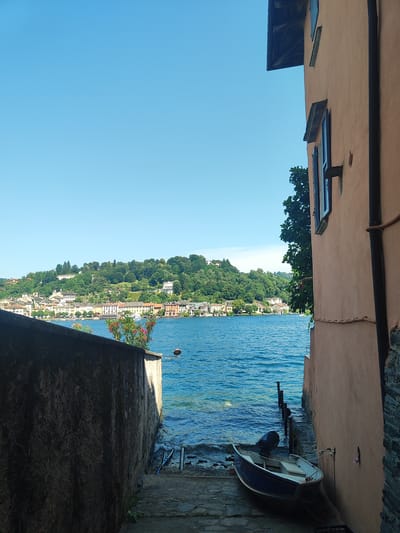 The image depicts a view of the lake in Orta San Giulio, Italy. The foreground features a weathered stone wall on the left and a peach-colored building on the right, with a narrow stone path leading down to the water. A small blue and white boat with a black outboard motor is resting on the stone path, partially in the water. In the middle ground, the vibrant blue lake stretches out, with a picturesque town lining the opposite shore. The town is characterized by buildings with colorful facades, nestled against a backdrop of lush green hills dotted with trees and houses. The sky above is a clear, brilliant blue. The scene conveys a sense of tranquility and a typical summer day by the lake.