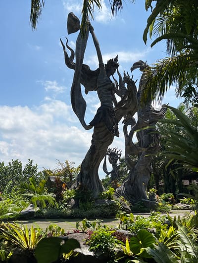 The image depicts a sculpted stone statue of a female figure, possibly a deity or mythological character, in a lush, tropical setting. The statue appears to be part of a landscaped garden, with a variety of plants and trees surrounding it. The sky is visible in the background, indicating an outdoor location. The statue is likely located in Kedewatan, Indonesia.