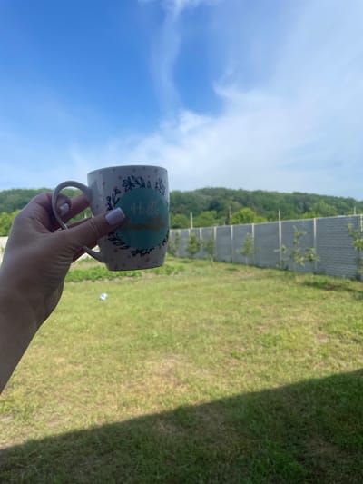 The image captures an outdoor scene in Petrivka, Ukraine, on a bright day. In the foreground, a left human hand, adorned with light lavender nail polish, holds a white ceramic mug. The mug features pink polka dots and a central teal circular design with green leaf motifs, containing the golden cursive text "Hello Beautiful." The ground beneath consists of a somewhat patchy green and dry brown grassy area, with a distinct dark shadow extending diagonally from the lower right corner. In the mid-ground, a lengthy, segmented grey concrete fence runs horizontally, behind which small green saplings are planted at intervals. The background reveals a continuous line of green forested hills under a clear blue sky dotted with soft white clouds, more concentrated towards the upper right.