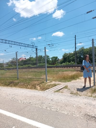 The image depicts a view of a railway track in Krymsk, Russia, under a clear blue sky with scattered white clouds. A young woman, wearing a light blue dress and carrying a dark bag over her shoulder, stands near a fence. She is looking to the right, away from the camera. The fence, made of metal mesh with grey posts, runs along the railway. Beyond the fence, railway tracks are visible, with sparse vegetation and trees in the background, including a house with a red roof. A road with a white line marking is in the foreground. The overall scene suggests a sunny day in a semi-urban or rural setting near a railway line.