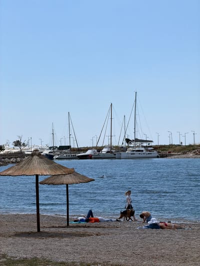The image shows a beach scene in Glifada, Greece. Along the shoreline, several sailboats are docked in a marina. Two straw umbrellas stand on the beach near the water's edge. People are present on the beach; a woman is walking two dogs along the shore. Other beachgoers are relaxing on the sand. The sky is clear and blue.