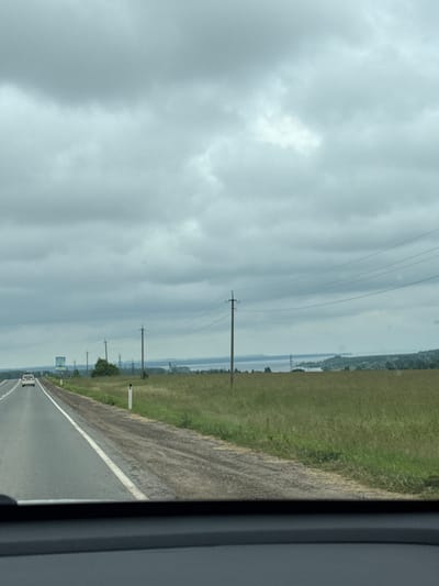 The image shows a view from inside a vehicle traveling on a highway. The road has a white dividing line and a shoulder. On the right side of the road, there is a grassy field with scattered telephone poles and power lines. In the distance, there is a body of water and some industrial structures. The sky is overcast with dark clouds. A white car is visible ahead on the road. A billboard is visible on the left side of the road. The scene appears to be a rural or semi-rural area in Russia, possibly on the outskirts of Kryukovo, a microdistrict in the Zelenograd Administrative Okrug of Moscow, Russia.