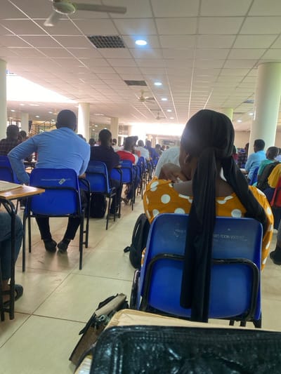 This image captures an indoor scene depicting a large room, likely a classroom, examination hall, or study area, occupied by numerous individuals. The perspective is from behind a few people in the foreground, looking down rows of seated attendees.
The setting features a bright white grid ceiling with evenly spaced recessed lighting and several ceiling fans, suggesting a well-lit and possibly air-conditioned environment. Large, light-colored pillars are visible, indicating a substantial architectural space.
In the immediate foreground on the right, a person wearing a black head covering and a yellow garment with white polka dots is seated on a blue chair, facing away from the camera. A black backpack is on the floor beside them. To the left foreground, another individual in a light blue long-sleeved shirt is seated at a blue chair with a wooden tablet arm. Further into the room, many more people, mostly men, are seen from behind, occupying similar blue chairs arranged in orderly rows.
The activity appears to be quiet and focused, suggesting the individuals are engaged in a lecture, taking an examination, or studying. Their posture implies concentration on something in front of them.
Notable details include the blue plastic chairs with black metal frames. White text is visible on the backs of some chairs; for instance, "07/XCH/15390" is clearly legible on a chair in the foreground. Another chair further back shows "VIAGI FURNITURE" and "WWW.VIAGIFURNITURE.COM," identifying the furniture manufacturer. These details, along with the structured seating, reinforce the impression of an institutional or educational facility. The bright, uniform lighting suggests a daytime setting within this facility in Ihumwa, Tanzania.