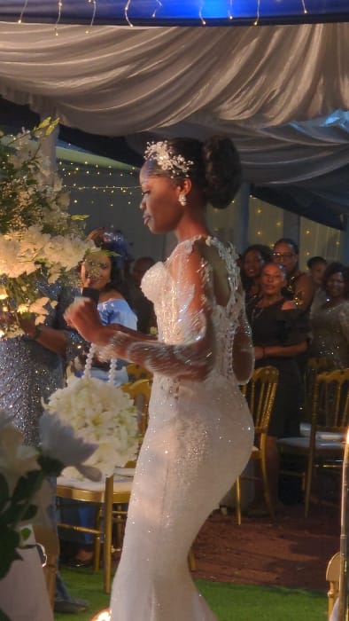A woman in a fitted, sparkling white gown adorned with lace poses, holding a bouquet of white flowers. Her hair is elegantly styled, accented with a hairpiece. Behind her, a group of people stands partially visible. The backdrop features draped white fabric and twinkling lights, creating a wedding scene. A blue pillar stands to the left.