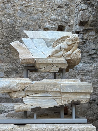 This image captures two fragmented ancient marble pieces, likely reliefs or architectural elements, displayed against a rugged, grey stone wall. The wall appears to be a natural rock face or an ancient construction. Modern metal support frames are partially visible, holding the heavy marble fragments in an upright position.

The fragment on the left features a sculpted figure, possibly a putto or Eros, with wings, depicted in a reclining or falling pose. The figure's head is turned to the right, with hands near its head. This marble piece is severely fractured, with several large cracks running through it. Below the sculpted figure, remnants of a Greek inscription are visible, including partial words like "ΣΟΝ ΑΙΚΑΒΕΣ" and "ΤΟΥΣ Ο".

The larger fragment on the right is a tall, rectangular marble slab, also broken into several pieces. It features deeply carved Greek capital letters. The inscription, spanning multiple lines, is clearer than that on the left fragment. Notable decipherable parts include:
- "...Ε[Π]ΙΜΟΝΔΙΟΙΣ ΤΩΝ..."
- "...ΠΡΩΙΗΣΩΝ ΤΗΜΕΤΕΡΩΝ..."
- "ΕΒΟΥΛΕΥΘΗ ΕΝ ΥΠΕΡΒΑΛΗΝ ΗΥΔΙΩΤΩΝ" (meaning "it was decided in Hyperbalos of the private citizens/residents")
- "...ΕΦΙΩΝ ΑΓΑΘΟΥ..." (likely part of "decree... of good")
- "...ΛΕΟΥΛΕ..."
- "...ΩΝ"

The artifacts are presented without specific lighting or time-of-day cues, suggesting an indoor or sheltered outdoor museum display setting. Given the location context of Kumluca, Turkey, these fragments are likely from the ancient city of Olympos.