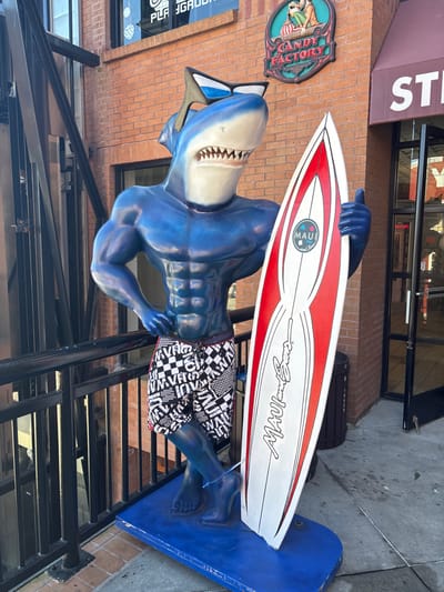A blue, muscular, anthropomorphic shark statue stands on a blue platform on a gray sidewalk, located outside a brick building on Cannery Row, Monterey, California, United States of America. The statue's head is white with visible sharp teeth and black sunglasses resting atop. It wears black and white checkered board shorts and holds a white surfboard with red accents and "MAUI and Sons" branding in its left hand, displaying a thumbs-up gesture. The statue is positioned against a black metal railing. On the reddish-brown brick facade behind it, a round green "Candy Factory" sign is mounted. To the right, a glass storefront door is visible, partially obscured by a dark trash can, with signage above reading "STE".