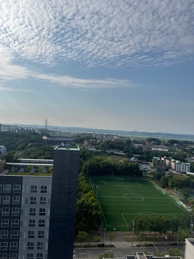 This is a vertical, wide-angle aerial shot of a suburban landscape in Pyeongtaek-si, South Korea, bathed in the soft light of a daytime sky. The image is divided horizontally by the horizon, with the upper half dominated by a vast expanse of blue sky filled with a dense pattern of small, puffy white clouds, creating a textured, almost quilted effect.
The lower half of the image reveals a sprawling urban and natural scene. In the foreground, a modern, multi-story building with a dark, textured facade and numerous windows dominates the right side. Adjacent to it is another residential building with a gray exterior and repeating window patterns. To the left of these buildings, a vibrant green soccer field with white markings, including a large circle and penalty areas, is clearly visible, bordered by trees and a fence.
Further into the landscape, a dense canopy of lush green trees covers hillsides and surrounds scattered residential buildings of varying sizes and architectural styles. The buildings are a mix of lighter-colored apartment blocks and smaller, more traditional houses. A wide, paved road runs alongside the soccer field, with parked cars visible. In the distance, a river or large body of water is visible, with more hills and a hazy skyline suggesting a flat plain stretching beyond. A prominent electrical tower stands tall amidst the greenery. The overall impression is one of a developed but verdant area under a clear, cloudy sky, likely taken from a high vantage point such as a tall building or drone. The time of day appears to be morning or late afternoon due to the soft lighting. No specific event or activity is clearly discernible, other than the presence of a sports field indicating potential recreational use.