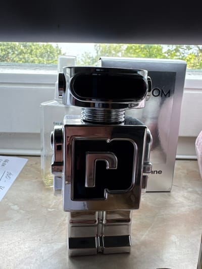 The image shows a robot-shaped perfume bottle placed on a marble countertop. Behind the bottle, a silver box with the text "LOOM" is visible. The background reveals a windowsill with trees and foliage outside. A small piece of paper with handwritten notes is also present on the countertop to the left of the bottle. The overall setting appears to be indoors, near a window. No specific location in Görlitz, Germany, is discernible from the image.