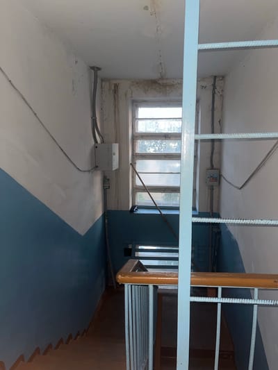 A first-person perspective looking down a stairwell in an apartment building, likely in Gorodets, Russia. The walls are painted in two tones: white on the upper half and blue on the lower half, with a sharp diagonal line dividing them. A light blue metal railing with a wooden handrail runs along the right side of the stairs. The stairs themselves are made of wood and appear worn.
Natural light streams in from a large window at the top of the stairwell, which is divided into multiple panes and appears to be slightly grimy. Electrical conduits and junction boxes are visible on the walls near the window. A light blue ladder-like structure runs vertically through the center of the frame, obscuring some of the view down the stairwell. The overall environment appears utilitarian and somewhat neglected. There are no people visible in the image.