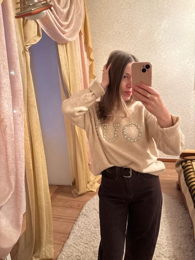 A mirror selfie captures a young woman standing indoors, holding a light-colored smartphone to take her picture. The image is oriented sideways, with the woman's head on the left and her feet extending to the right.
The woman has long, straight light brown hair and a neutral expression, looking directly into the phone's camera reflection. Her face is partially obscured by the phone. She is wearing a soft, light beige or cream-colored knitted sweater with ribbed cuffs. Prominently displayed on the front of the sweater are large, decorative letters formed by white pearls or beads. The letters "GU" are clearly visible, followed by what appears to be a "C" or "O," suggesting a brand name like "GUCCI." She is also wearing dark brown or black trousers, secured with a brown belt featuring a silver-toned buckle. A golden ring is visible on the ring finger of her right hand, which is holding the phone.
The background behind her offers glimpses of various indoor elements. To her left (top of the image when viewed sideways), there's a light-colored wall covered in textured wallpaper with a subtle, swirling or leafy pattern. To her right (bottom of the image when viewed sideways), the floor consists of light wooden planks, partially covered by a fluffy, off-white or cream-colored shaggy rug.
On the far left side of the image (bottom when viewed sideways), there are multiple layers of curtains. The topmost layer visible is a gold or yellow crushed fabric, beneath which is another layer of pinkish, possibly velvet or crushed fabric. A gap between the curtains reveals a light blue or lavender wall or surface. In the bottom left corner, a metallic object with a circular design and a reddish accent is visible, possibly part of a light fixture or decor. In the upper right corner (far right when viewed sideways), part of a piece of furniture, possibly a bed or bench, is visible, draped with a striped blanket in shades of brown and beige. The lighting appears to be ambient indoor light.