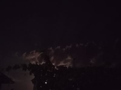 This is a nighttime image, likely taken outdoors, with very dark conditions. The sky is predominantly black with some discernible clouds that are faintly illuminated, possibly by moonlight or distant artificial light. In the foreground, silhouetted against the slightly lighter sky, are dark shapes of trees and foliage. A portion of a structure, possibly a roof or building edge, is visible on the far left of the image. A faint light source, appearing as a small white dot, is present in the lower portion of the frame, near the center. The scene lacks clear details of people or specific urban features due to the low light conditions. It is not possible to determine the exact location within Ucrainca, Moldova, based solely on this image.