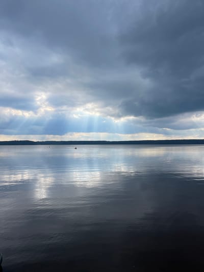 This media file depicts a serene natural landscape, likely a lake or large body of water, with a striking contrast between the sky and the water. The image is oriented vertically.

The left side of the frame is dominated by a dramatic sky. Dark, heavy clouds occupy the upper portion, with shafts of sunlight breaking through in the middle, creating a sunbeam effect. The sunbeams illuminate patches of lighter clouds and the blue sky beyond.

The right side of the frame shows the reflective surface of the water. The water appears calm, with ripples catching the light, creating streaks of white and gray that mirror the sky. The far horizon is marked by a dark, tree-lined silhouette, suggesting a forested shoreline.

There are a few small, distant objects on the water that could be boats or birds, with one object appearing to be a small red and white boat or sail. The overall mood is peaceful and contemplative, with the interplay of light and shadow creating a sense of depth and natural beauty. The time of day appears to be late afternoon or early evening, given the dramatic lighting and potential for sunsets, with the weather transitioning from cloudy to clearing. The location context places this scene in Vaskelovo, Russia. There is no visible text in the image.