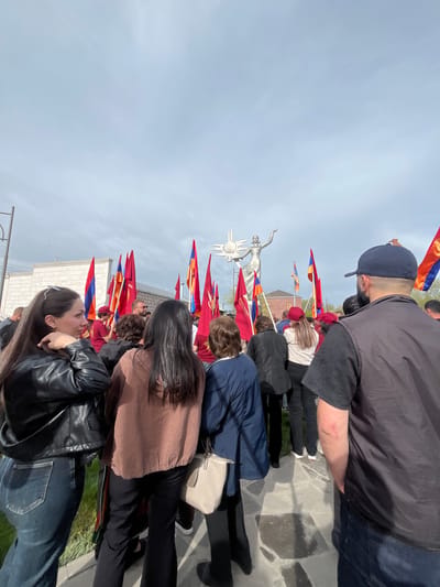 A horizontal eye-level shot depicts a crowd of people gathered in Akhuryan, Armenia. In the center of the crowd is a statue of a woman in a silver finish. The statue is surrounded by several flags of Armenia. People are wearing casual clothes, including jackets, caps, and casual tops. The weather is clear and sunny, and the sky is mostly cloudless. The location is urban, as evidenced by sidewalks and low buildings in the background.