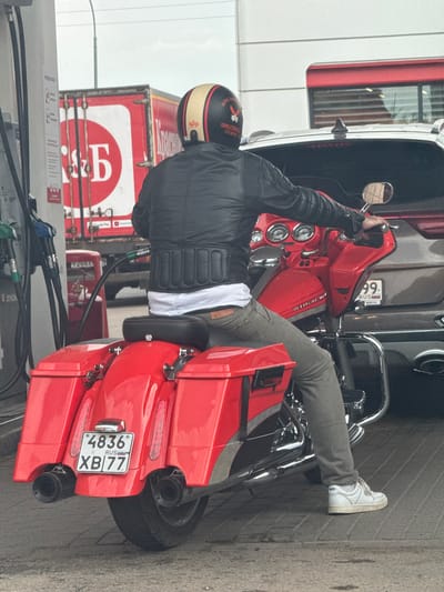 A person wearing a black leather jacket, a white t-shirt, grey pants, and white sneakers is sitting on a bright red motorcycle at a gas station. The motorcycle has two large red saddlebags on the back and chrome accents. The person is wearing a black and gold helmet with a red stripe. A fuel hose from a gas pump is inserted into the motorcycle's fuel tank. In the background, a red truck with a white logo featuring a stylized "Б" is visible. The license plate on the motorcycle reads "4836 XB 77 RUS." The scene appears to be a roadside gas station in or near the city of Ermolino, Russia, indicated by the license plate format and truck branding.