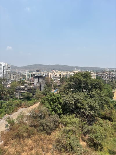 The image depicts a cityscape from an elevated viewpoint in Ambernath, India. A mix of low-rise and high-rise buildings are visible against a backdrop of rolling hills under a hazy sky. Lush greenery and dense trees flank the built environment on the right side of the image, contrasting with a dry, overgrown hillside. The buildings appear to be a mix of residential and commercial structures. The terrain slopes downward towards the bottom of the frame, exposing a pathway or clearing littered with debris. The time of day appears to be daytime, given the lighting conditions. No people are clearly visible in the scene.