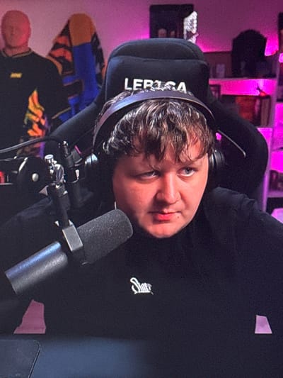 A young man with curly brown hair, wearing a black headset and a black shirt with a white logo on the chest, is seated in a black gaming chair. He is looking directly at the camera with a neutral expression. A microphone is positioned in front of him, and his hands are not visible. The background is out of focus but appears to be a room with shelves filled with objects and pink lighting. There are also two mannequins in the background, one on the left wearing a black shirt with yellow accents and another partially visible behind him wearing a colorful garment. There is no indication of the specific location in Gothenburg, Sweden.