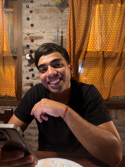 A young adult male is seated at a dark brown table indoors, located in the city of Partido de Quilmes, Argentina. He is looking directly at the camera with a wide smile, displaying his teeth. He has dark hair, styled to the side, and a small stud piercing in his left nostril. He wears a black, short-sleeved t-shirt. A red string bracelet is visible on his right wrist. His left hand is holding a gray smartphone, which rests on the table alongside a partially visible plate with an abstract design. The background consists of an exposed brick wall, adorned with a vertical string of multi-colored, reflective spherical ornaments. Orange polka-dotted sheer curtains are draped over two windows flanking the male subject. A decorative object with blue and yellow components hangs above his head. The scene is illuminated by warm, artificial light.