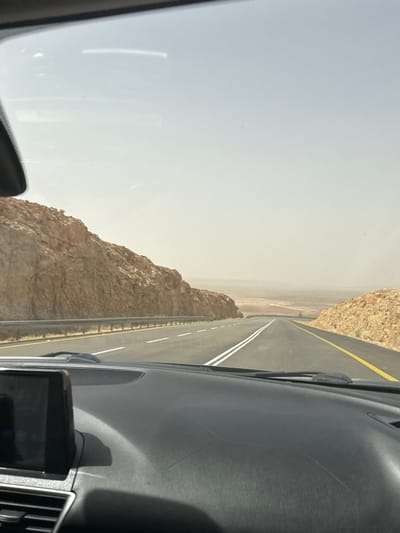 The image shows a view from inside a vehicle traveling along a desert highway in Ramat Negev Regional Council, Israel. The road is paved with white and yellow lines indicating lanes. On either side of the road, there are rocky cliffs with sparse vegetation. The sky is hazy and overcast. The vehicle's dashboard is visible in the foreground, featuring a navigation device. The scene suggests a remote and arid landscape.