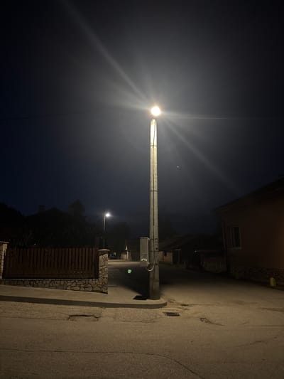 The image depicts a street in Dimovo, Bulgaria at night, illuminated by a tall, concrete lamppost with a bright light at its apex, casting rays across the dark sky and reflecting on the wet asphalt. The lamppost is situated on the right side of the street, near a drainage grate. To the left of the lamppost, a wooden fence with stone pillars lines a sidewalk. In the background, silhouetted houses and trees are visible against the twilight sky. The street itself is paved and appears to have some minor damage, including potholes. The overall atmosphere is quiet and still, characteristic of a rural town at night.