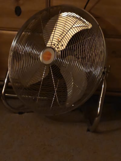 A metallic, oscillating fan is positioned on a carpeted floor, situated in front of a wooden dresser. The fan is turned on, as indicated by the light emanating from its center and the slight blur of the blades. The lighting in the room is dim, casting shadows. The scene is located in Novaya Polyana, Russia.