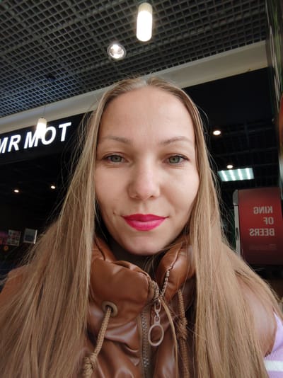 A medium shot captures a woman with long, blonde hair and striking green eyes. She is looking directly at the camera, with a subtle smile playing on her lips, enhanced by vibrant pink lipstick. She's wearing a brown leather jacket over a light purple and white striped garment. The background suggests an indoor setting, possibly a shopping mall or a commercial area.
Above her, a dark ceiling grid with square openings is visible, punctuated by a few bright lights. To her left, a prominent white sign with the text "MR MOT" is illuminated against a dark background. To her right, a vertical red sign displays white text which appears to read "KING OF BEERS" in a mirrored fashion, implying it might be a reflection or a sign meant to be viewed from the other side.
The lighting suggests it is daytime, but the indoor setting makes it difficult to determine the exact time or weather conditions. The woman's expression is neutral and confident. The overall impression is that of a casual selfie taken in a public, commercial space. The location context of Syktyvkar, Russia, adds a geographical anchor to the scene.