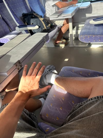The image depicts the interior of a train carriage, likely during the daytime given the strong sunlight casting shadows. The seats are upholstered in a purple fabric with a patterned design of diagonal white and yellow lines. In the foreground, a person's outstretched hand is visible, with their arm and leg also in frame. This person is wearing a grey denim skirt and white socks with grey sneakers. Another person's legs and feet are visible further down the aisle, wearing white shorts and sandals. A backpack rests on a seat to the left. The sunlight streaming through the windows creates bright patches on the floor and seats, suggesting a clear day. The scene appears to be a normal commute or journey on a train. No visible text is present in the image.