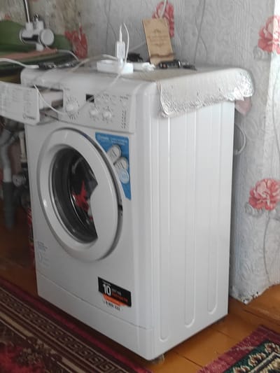 A white washing machine, with a transparent lid and a pink floral patterned fabric draped over its top, is situated on a wooden floor next to a patterned rug. Several white electrical cords and a white charging block are connected to the top of the machine. The washing machine has a blue and white sticker on its front door, which appears to be a product label. The background consists of a textured, light-colored wall with a floral pattern. To the left of the washing machine, a white faucet with a silver handle is mounted on a wall. The scene suggests an indoor domestic setting. No people are visible in the image. The image appears to have been taken in Некрасовка, Kazakhstan.
