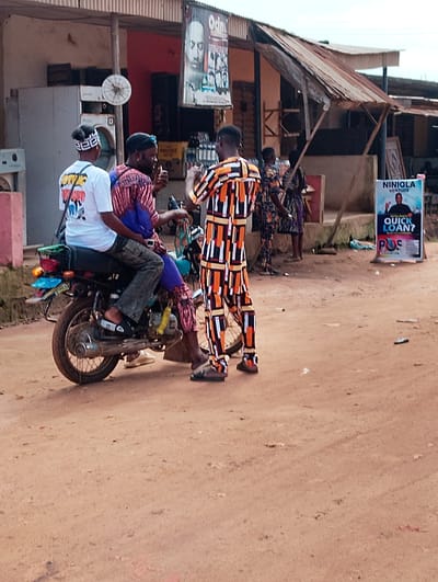 In Orudu-Olokan, Nigeria, three individuals are gathered near a motorcycle parked on a dirt road. One person, wearing a white t-shirt with black lettering and a white patterned headband, sits at the rear of the motorcycle. Another individual, dressed in a patterned blue and red garment, sits behind the driver, who is wearing a colorful orange, black, and white patterned outfit and black sunglasses. The driver appears to be speaking or gesturing with his left hand, holding a small object.
Behind the individuals, a weathered building with a corrugated metal roof stands. A sign advertising "NINIOLA VENTURE" with the tagline "QUICK LOAN?" and the logo "POS" is prominently displayed. The environment is dusty and appears to be an outdoor market or roadside commercial area, with other buildings and signs visible in the background. The overall scene suggests a casual interaction or transaction taking place in a bustling, informal setting.