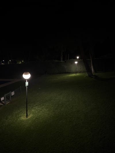 This image depicts a nighttime scene in a green area, possibly a park or garden, in El Escorial, Spain. Several lampposts with spherical globes illuminate the area with a warm light, casting pools of illumination on the well-maintained grass. A dark, dense hedge forms a backdrop behind the grass, with trees visible beyond it, their forms silhouetted against the night sky. In the distance, more lampposts can be seen, suggesting a public space. To the left of the frame, a portion of a swimming pool and a metal fence are visible, indicating the presence of recreational facilities. The overall atmosphere is serene and quiet, with no people or animals present. The lighting suggests that it is a safe and accessible area even after dark.