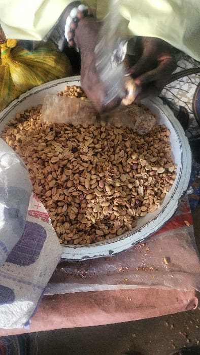 A high-angle, close-up shot captures a vendor activity in Fagge D1, Nigeria. The central subject is a large, circular, white enamel basin with a worn blue rim, filled to capacity with light brown, shelled peanuts; some retain reddish skin fragments. Above the basin, two dark-skinned hands are actively engaged with the peanuts. The left hand grasps a transparent plastic bag containing peanuts, while the right hand is positioned within the basin, appearing to scoop or manipulate the peanuts, possibly into another plastic bag. Small white, spherical objects are visible on the fingers of the left hand. The immediate environment includes a light yellow fabric visible at the top, a bright yellow bundled object to the upper left, and a dark object with a blue pattern to the upper right. To the lower left, a portion of a white woven sack with purple markings is visible. The surface beneath the basin is reddish-brown, partially covered by clear plastic sheeting, with scattered peanut fragments.