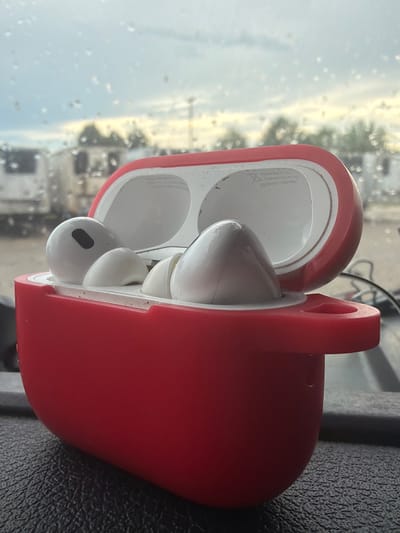 The image shows a close-up, eye-level view of an open red charging case for wireless earbuds. The earbuds themselves are white and are partially visible within the case. The charging case is positioned on a dark, textured surface, possibly a dashboard or a car interior. In the background, a window shows a blurry outdoor scene with trees, vehicles, and a cloudy sky with visible raindrops on the glass. The overall impression is a still life of personal electronic devices set against a backdrop suggesting travel or an outdoor environment in Murfreesboro, United States of America.