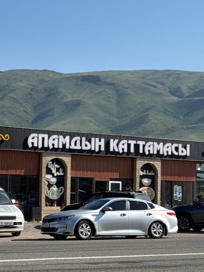 The image depicts a roadside scene in Sasyk-Bulak, Kyrgyzstan. A building with a large vertical sign reading "АПАМДЫН КАТТАМАСЫ" stands alongside a road. Cars are parked in front of the building. A mountain range is visible in the background. The sky is clear and blue.