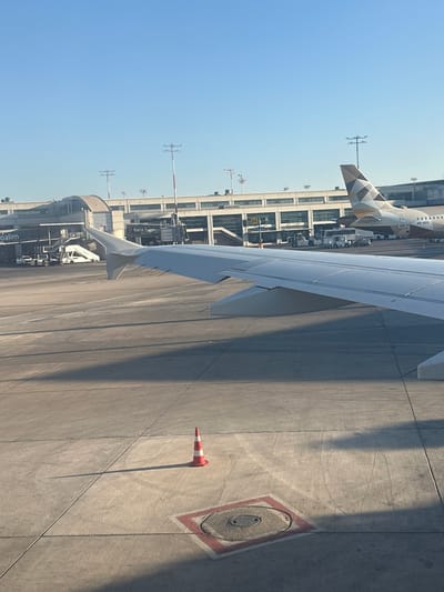 The image shows an airport tarmac on a clear, sunny day, likely in the morning or afternoon given the long shadows. The perspective is from inside an airplane, looking out over the wing and onto the tarmac. The scene is dominated by the concrete expanse of the tarmac, with the large wing of the airplane taking up a significant portion of the frame.
Several airport-related objects are visible. An airplane with a distinctive patterned tail, possibly belonging to Etihad Airways due to the triangular geometric design, is parked in the background. Various service vehicles and equipment are also present near the terminal building. A bright orange and white traffic cone lies on the tarmac, casting a long shadow. In the distance, the terminal building is visible, a long, multi-story structure with many windows. Airport lighting masts, with red and white markings, are also scattered across the tarmac.
There is visible text on the side of the terminal building, including "bank hap"}," which likely refers to Bank Hapoalim, a major Israeli bank. Another partial word, "oalim," is also visible, possibly part of the same building or another sign. The overall impression is one of a busy airport environment preparing for or just having completed a flight. The clear sky and strong sunlight suggest good weather conditions.