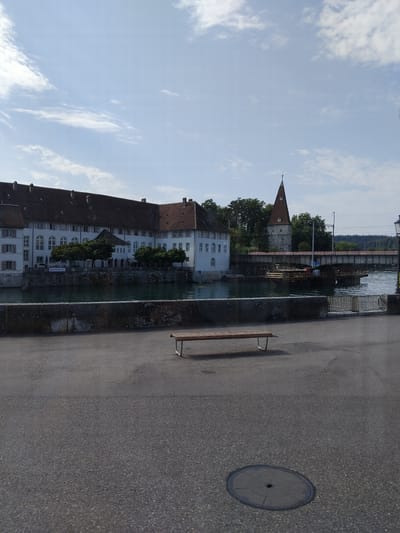 The image depicts a view of the Aare river in Solothurn, Switzerland, looking towards the historic hospital building (Altes Spital). The large, white, multi-story building with a red tiled roof dominates the left side of the frame. It features numerous windows, some with decorative red and white shutters, and a banner that reads "SCHWEIZERGARTEN". Trees line the riverbank in front of the building.
A tower with a distinctive reddish-brown conical roof stands to the right of the hospital building, suggesting a historical architectural style. A bridge, likely a railway bridge given the presence of tracks, spans the river in the background. Construction or renovation work is visible on the bridge, with scaffolding and wooden structures in the water. The river itself is a deep, murky green, reflecting the sky.
In the foreground, a grey asphalt path runs parallel to the river. A simple wooden bench with metal legs is positioned on this path, facing the river and the buildings. Further down the path, a circular metal manhole cover is visible. The overall environment suggests a public space along the river, likely a promenade or walkway, with elements of historical architecture, infrastructure, and recreational facilities. The sky is partly cloudy with a pale blue hue, indicating a daytime scene.