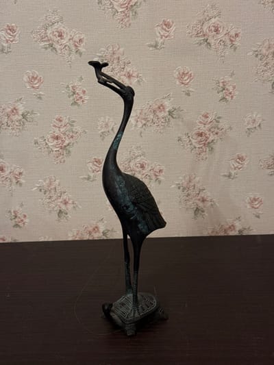 The scene is located in the city of Zelenogradskiy, Russia. A dark-colored statuette, approximately 30 cm tall, depicting a stylized crane standing on the back of a tortoise, is centrally positioned on a dark brown, smooth horizontal surface. The crane, rendered with an elongated neck and detailed feathers, exhibits areas of blue-green patina suggesting aged bronze or a similar metallic composition. Its head is raised, holding an object resembling a stylized cloud or reishi mushroom in its beak area. The tortoise base is also dark, featuring segmented shell details. To the left of the statuette's base, a single, light-colored hair-like fiber rests on the dark surface. The background consists of a wall covered in beige or off-white wallpaper patterned with repeating clusters of light pink roses and green foliage.
