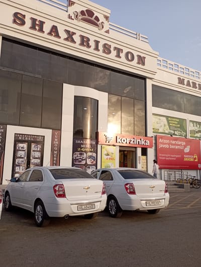 This media file captures a street scene in Termez, Uzbekistan, featuring a modern commercial building with prominent signage. The building displays large, stylized lettering that reads "SHAXRISTON" and "MARK." Below this, there are various advertisements, including one for "korzinka" (a supermarket or convenience store), another promoting furniture ("MEBEL") with offers like "BUYURTMA ASOSIDA MEBELLAR" (Furniture on order) and "Arzon narxlarga javob beramiz!" (We guarantee low prices!). One ad specifically mentions "SHAXRISTON MEBEL 2-QAVAT" (Shaxriston Furniture, 2nd floor).
Parked on the asphalt in front of the building are two white Chevrolet cars. The rear license plate of the closest car is visible and reads "75L 991YA." The second car's license plate is partially visible and reads "10L 684KB."
A person in a light-colored shirt is walking near the entrance of the building. The lighting suggests it is either early morning or late afternoon, with the sun casting long shadows and creating a warm, golden hue on the building's facade. The sky is clear and blue. The scene is one of everyday commercial activity in a city setting.