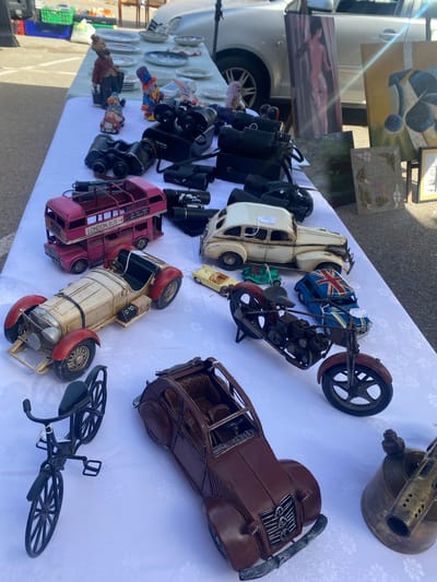 The image shows a white tablecloth laid out on a table at an outdoor market. The tablecloth is covered with various vintage and collectible items, primarily metal toy vehicles and some decorative figurines.

In the foreground, to the left, is a black metal toy bicycle with intricate detailing, including spokes on the wheels and pedals. To the right of the bicycle is a dark brown, classic-style toy car with a visible grill and chrome accents. Further to the right is a brass blowtorch.

Moving up the table, there is a red and white two-tiered toy London bus. Next to it is a cream-colored vintage toy car with red accents on the fenders and wheels. A small yellow toy car and a green toy car are visible behind it.

Further up the table, there are several pairs of black binoculars, some in cases. There are also some decorative ceramic clown figurines and other small figurines. In the background, on the table, are several plates and bowls. Behind the table, partially visible, is a silver car and some crates. To the right of the table, leaning against the car, are two framed artworks.

The overall impression is a display of vintage items for sale at a market. The market appears to be located in San Luis de Sabinillas, Spain, as suggested by the context. The lighting indicates it is daytime, likely outdoors. No people are prominently featured in the image.